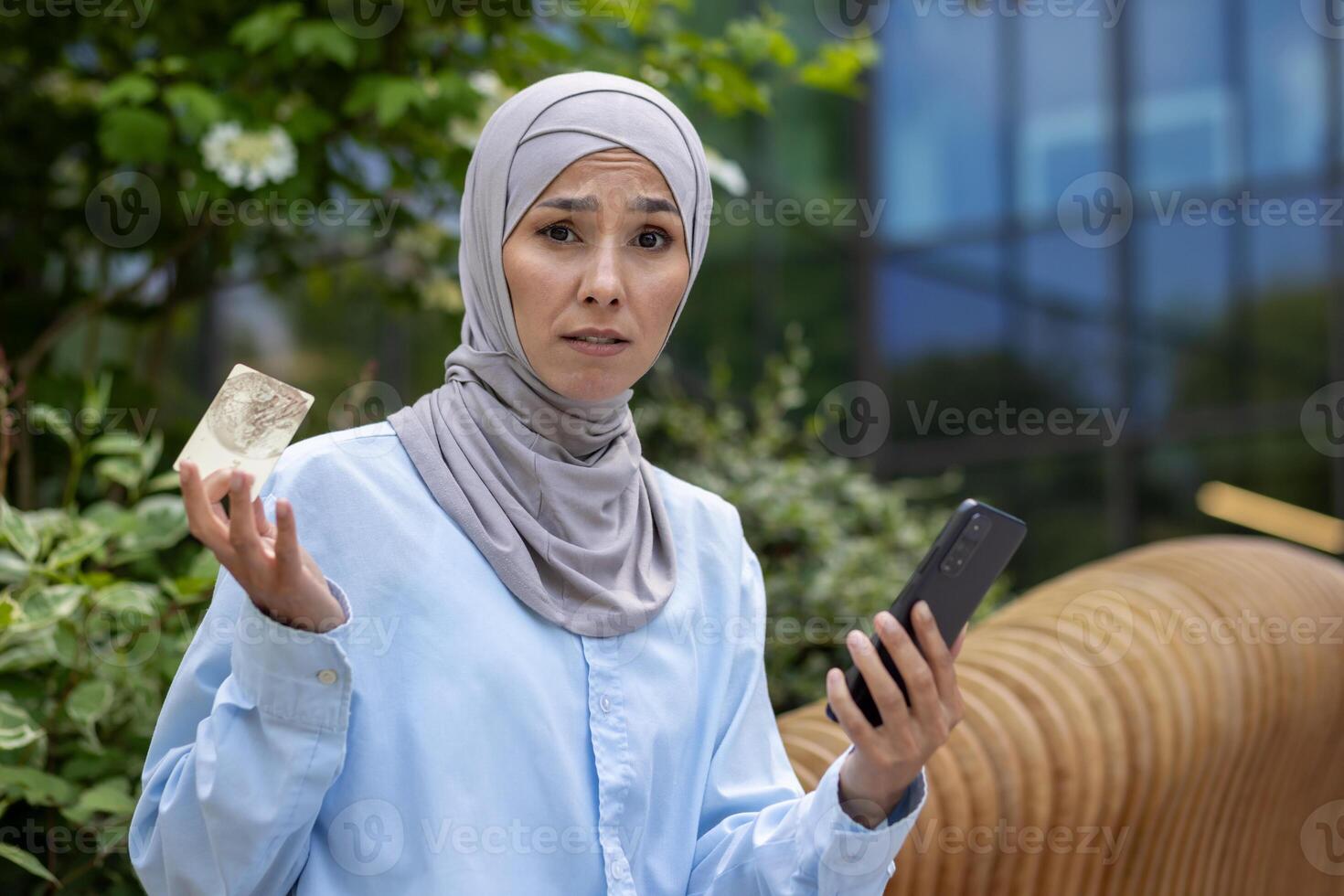 Upset woman in hijab looking at camera outside office building, business woman holding bank credit card and phone, rejected money transfer, and online fraud photo