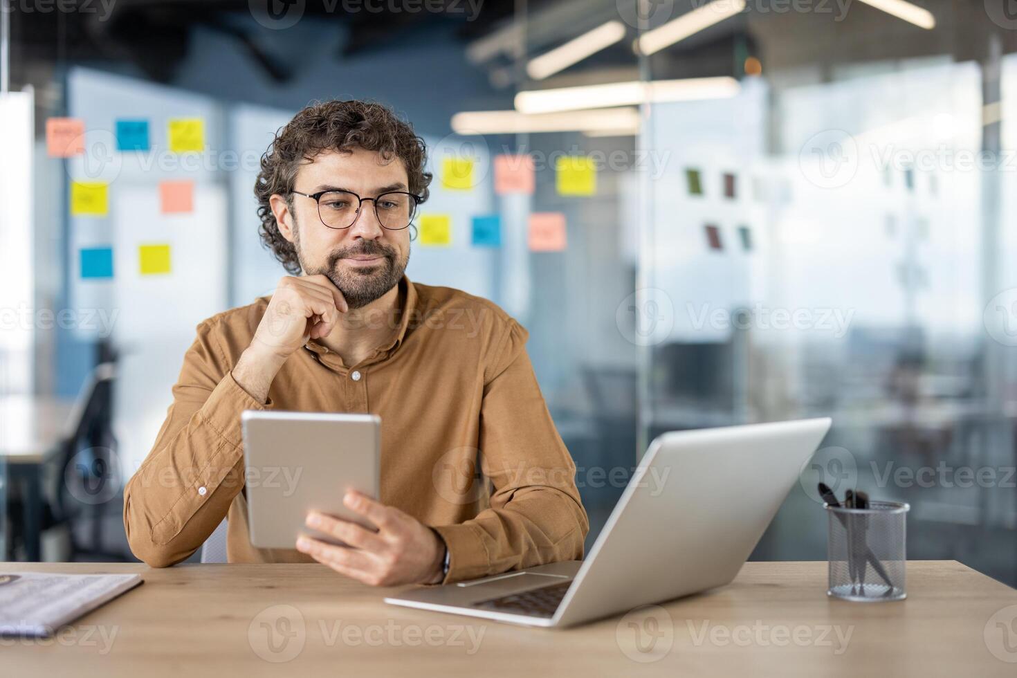 Focused professional man using tablet and laptop at a contemporary workspace with sticky notes in background. photo