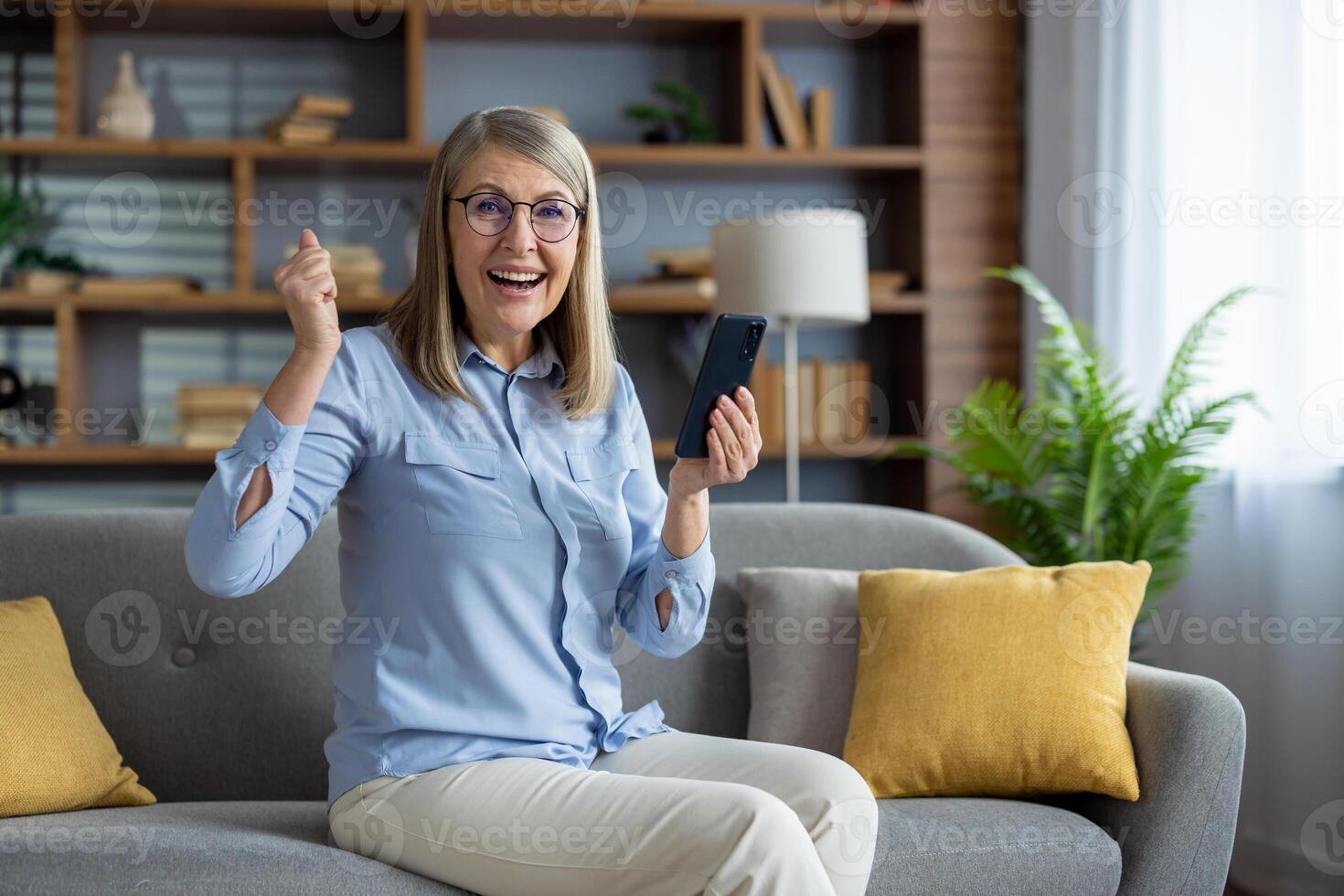 Joyful adult woman sitting on sofa, exulting with raised fist and looking at phone, expressing success and happiness indoors. photo