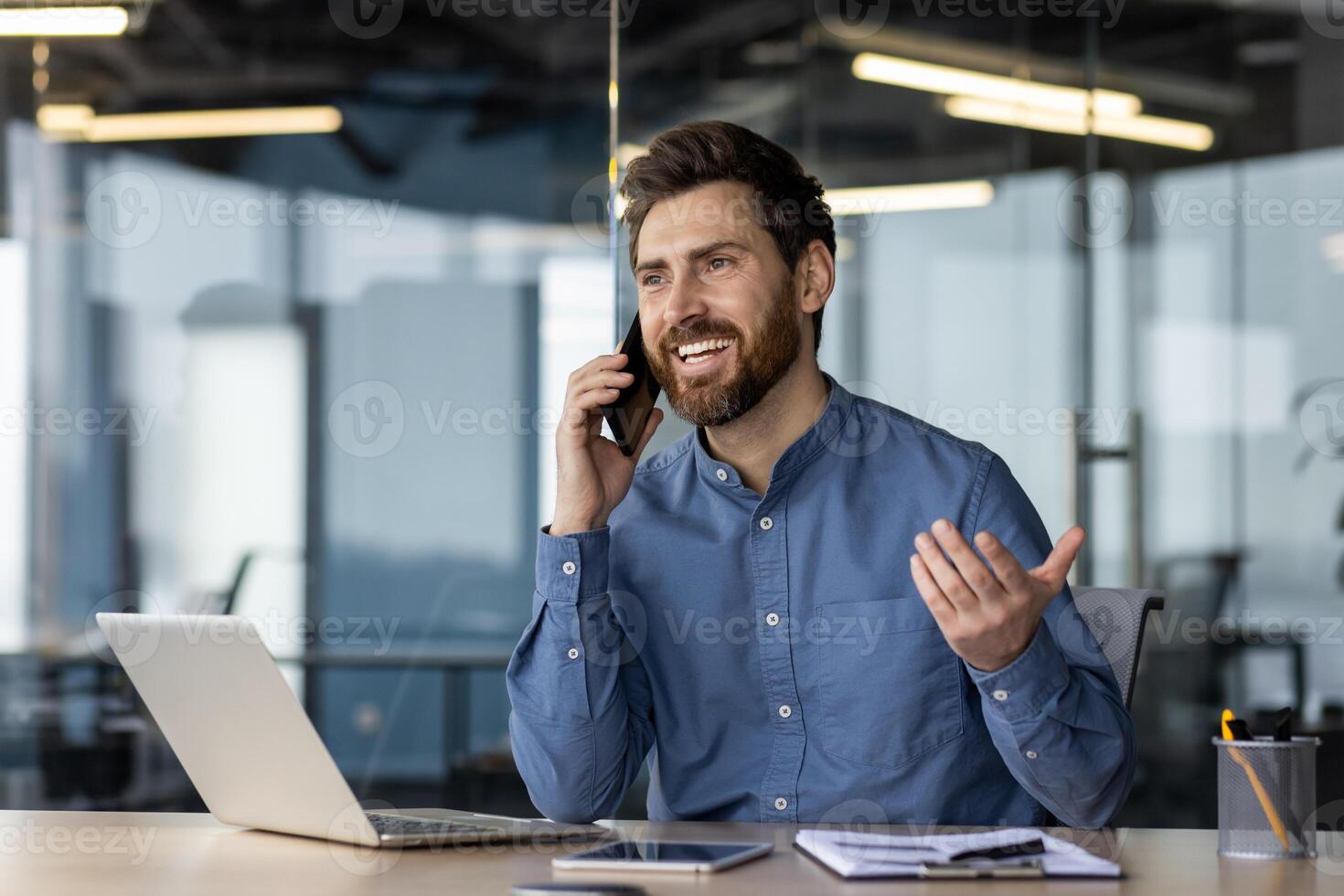 Smiling young man businessman has a business conversation with clients and partners on the phone, sits in the office at the table and explains actively gesturing with his hands. photo