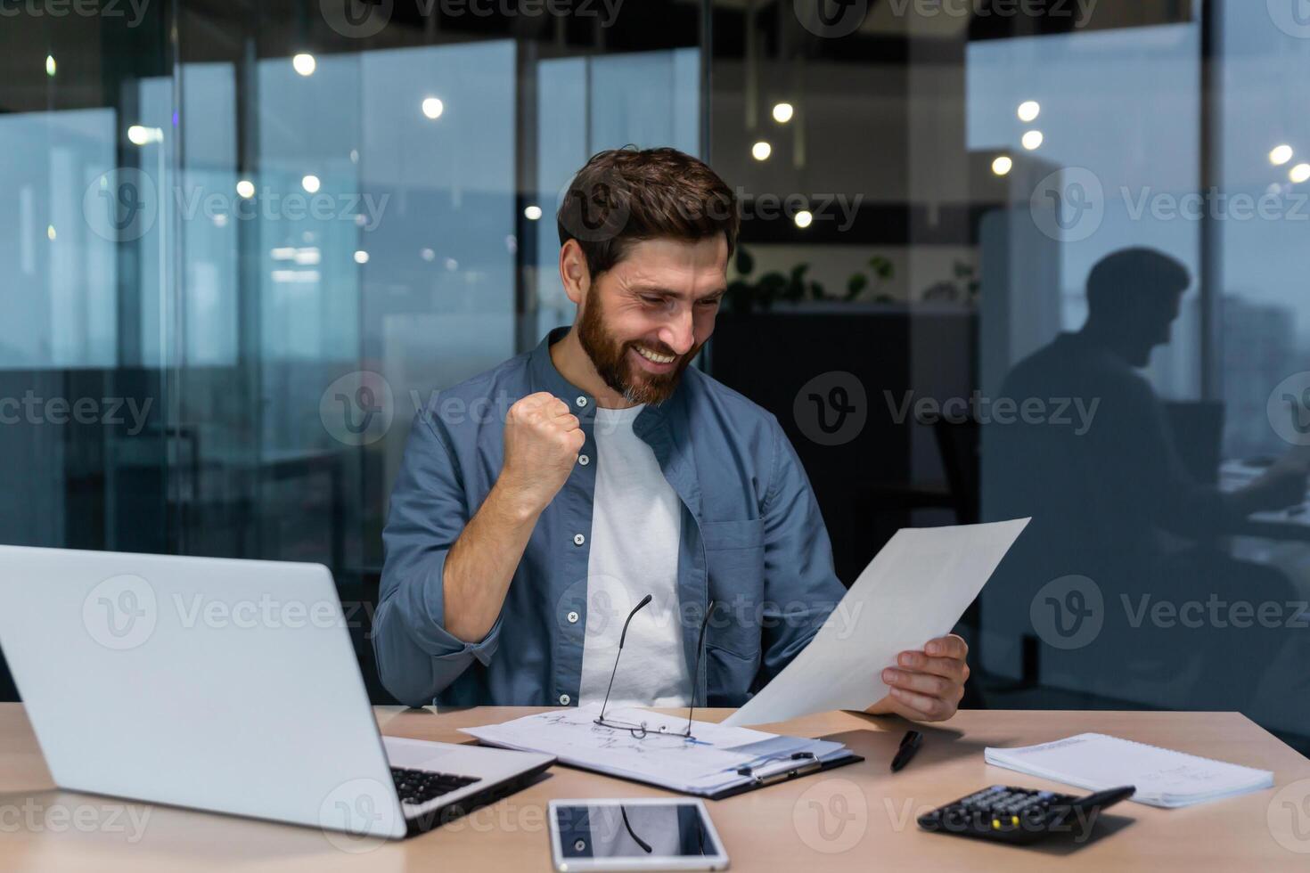 exitoso empresario en casual camisa haciendo papeleo, jefe con barba y lentes sentado a escritorio a lugar de trabajo utilizando ordenador portátil trabajando con documentos, participación mano arriba celebrando victoria y triunfo. foto