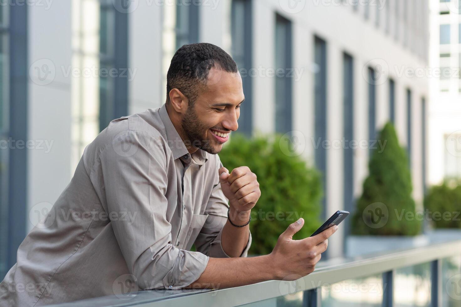 contento africano americano empresario mirando a teléfono inteligente pantalla, hombre en camisa fuera de moderno oficina edificio recibido bueno Noticias de ganar y éxito en línea, jefe celebrando financiero triunfo foto