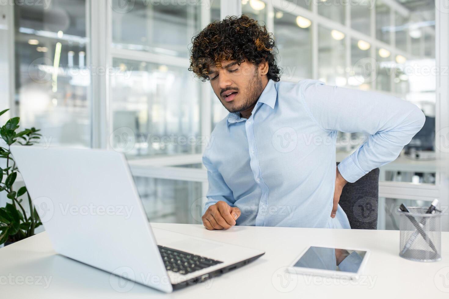 un joven masculino oficina trabajador sentado a su escritorio en un moderno oficina, experimentando espalda dolor mientras trabajando en su ordenador portátil. foto