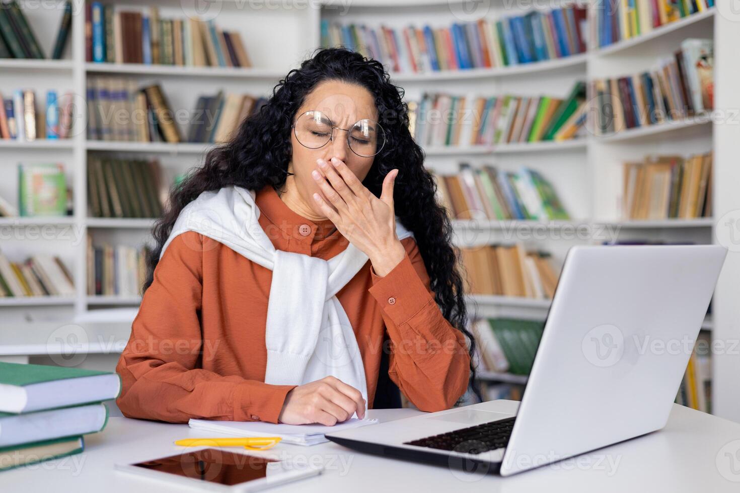 Sleepy and overtired young student studying in campus academic library until late, Hispanic woman preparing for entrance exams among books, using laptop to view online courses. photo