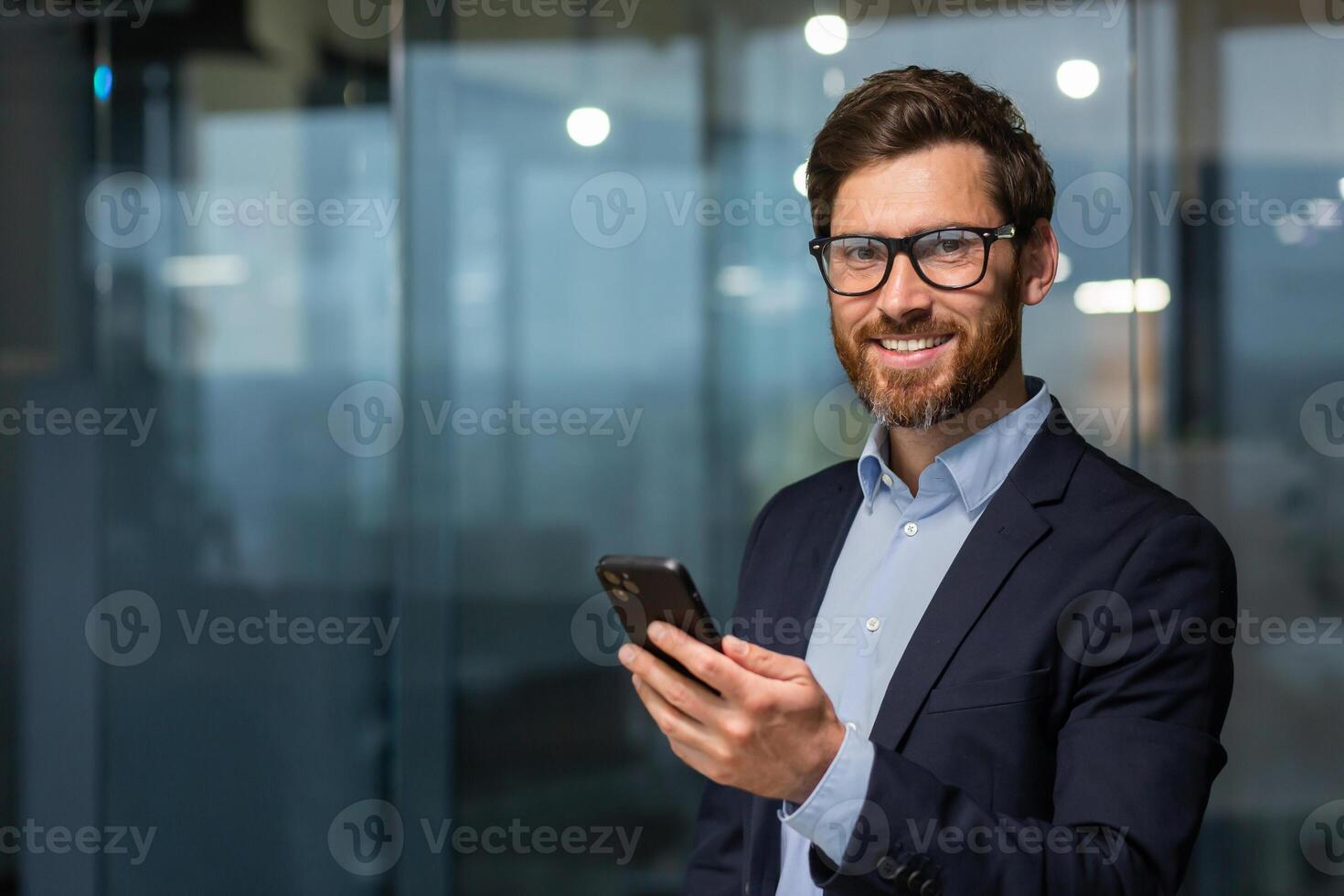 retrato de exitoso maduro empresario inversor, hombre en lentes y barba sonriente y mirando a cámara, jefe en traje a trabajo dentro oficina participación teléfono inteligente foto