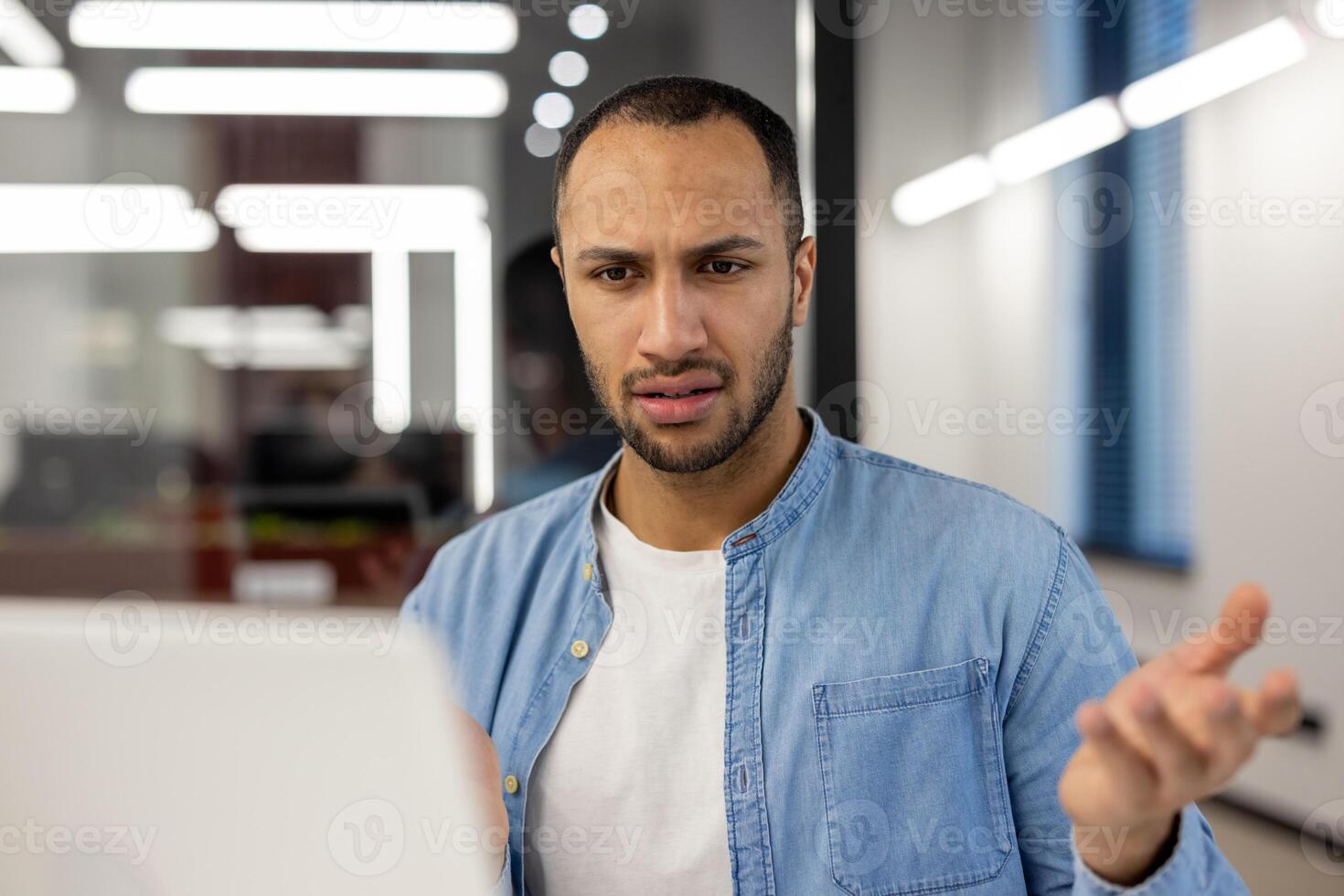A young male professional in a blue denim shirt appears perplexed and frustrated while working at his computer in a modern office setting, expressing a common workplace challenge. photo