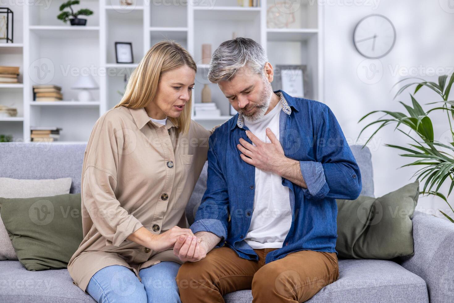 asustado esposa participación mano de marido teniendo corazón ataque y participación mano a pecho, sentado en sofá en vivo habitación a hogar. foto