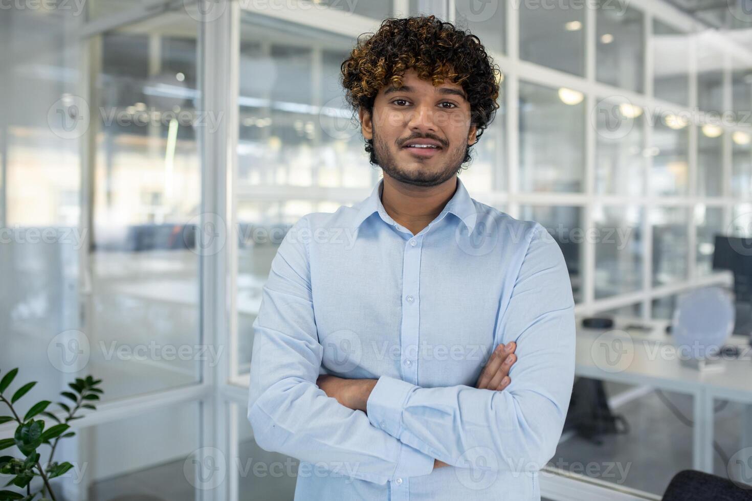 A portrait of a confident young businessman with curly hair, standing arms crossed in a light-filled, contemporary office environment. photo