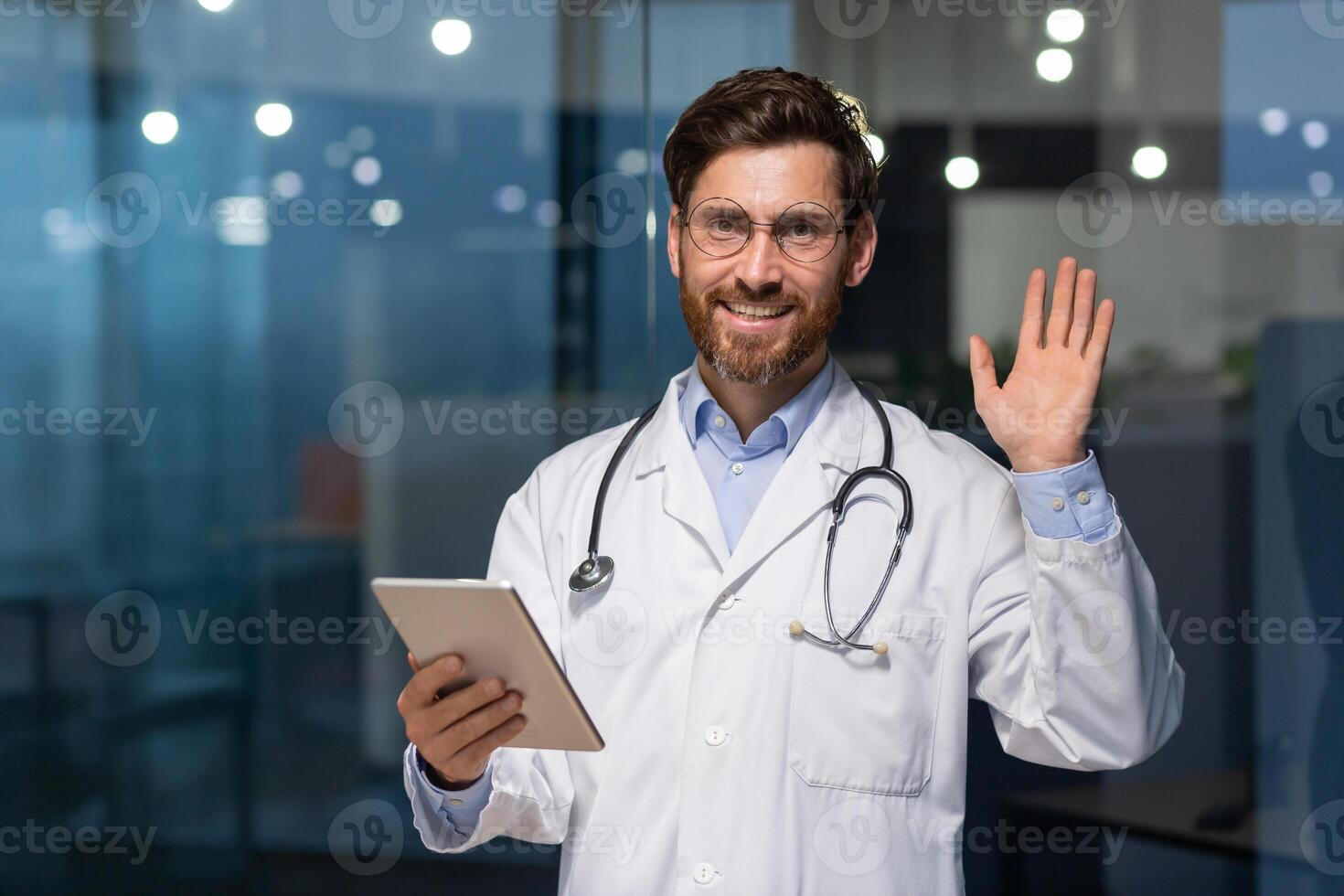 Senior mature doctor with tablet looking at camera and holding hand up greeting gesture, man in white medical coat working inside modern clinic, using tablet computer at work. photo