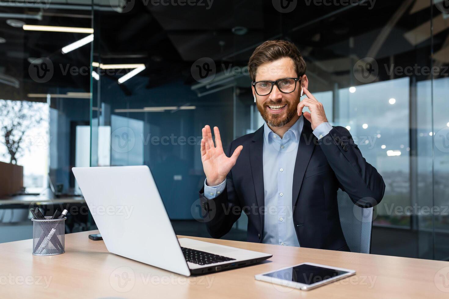 Portrait of a successful businessman inside a modern office, a man in a business suit uses earphones for a call and remote communication, an investor looks at the camera and uses a laptop. photo