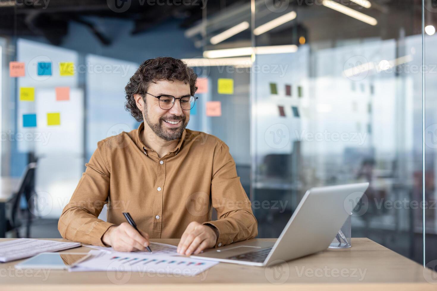 A cheerful business professional analyzes data on his laptop in a contemporary office setting. notes visible in the background suggest project planning. photo