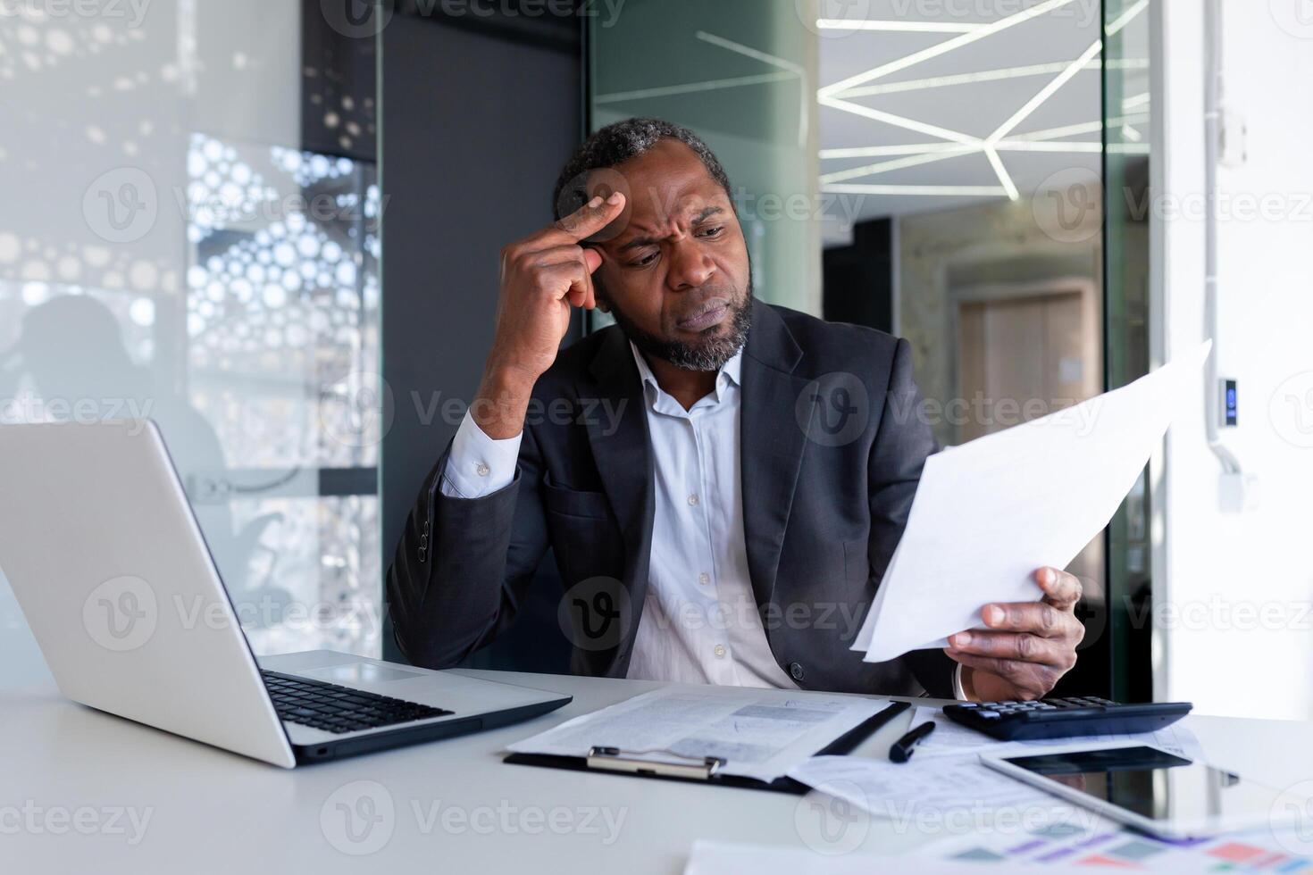 Serious thinking businessman behind paper work inside office, senior mature boss reviewing accounts contracts and financial reports, african american businessman solving accounting analytical tasks. photo