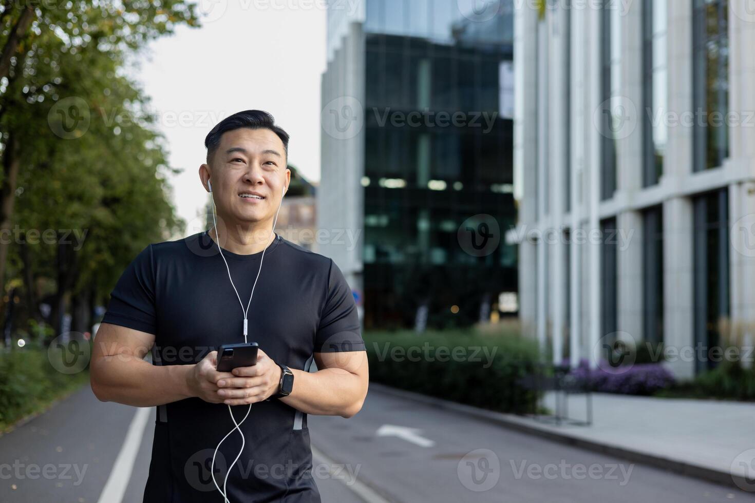 An Asian young smiling man is doing a morning jog on the city streets. Standing in sportswear and headphones, holding a phone. photo