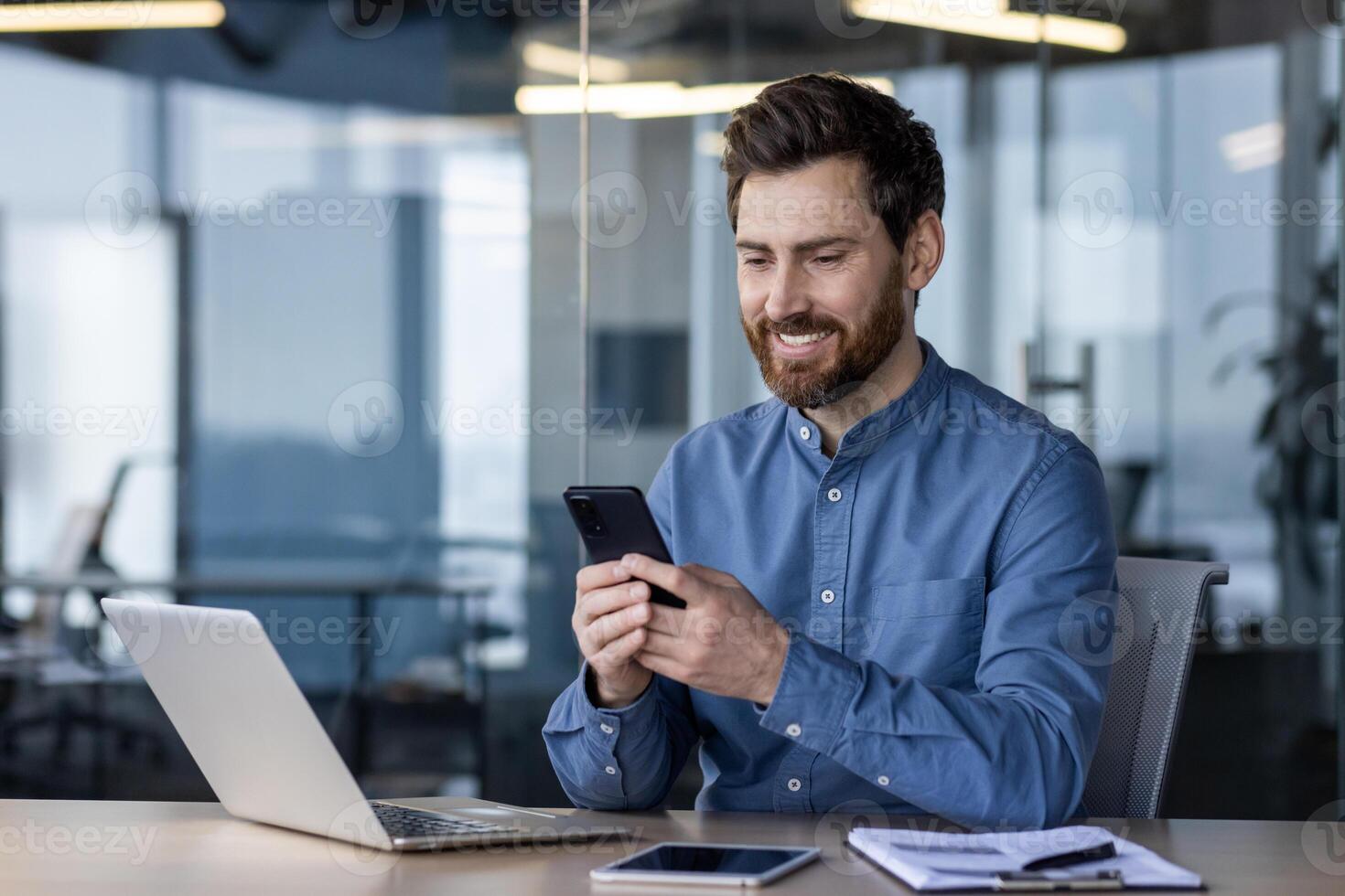 A smiling young male office worker sits in the office at a desk and uses a mobile phone, writes a message, corresponds. photo