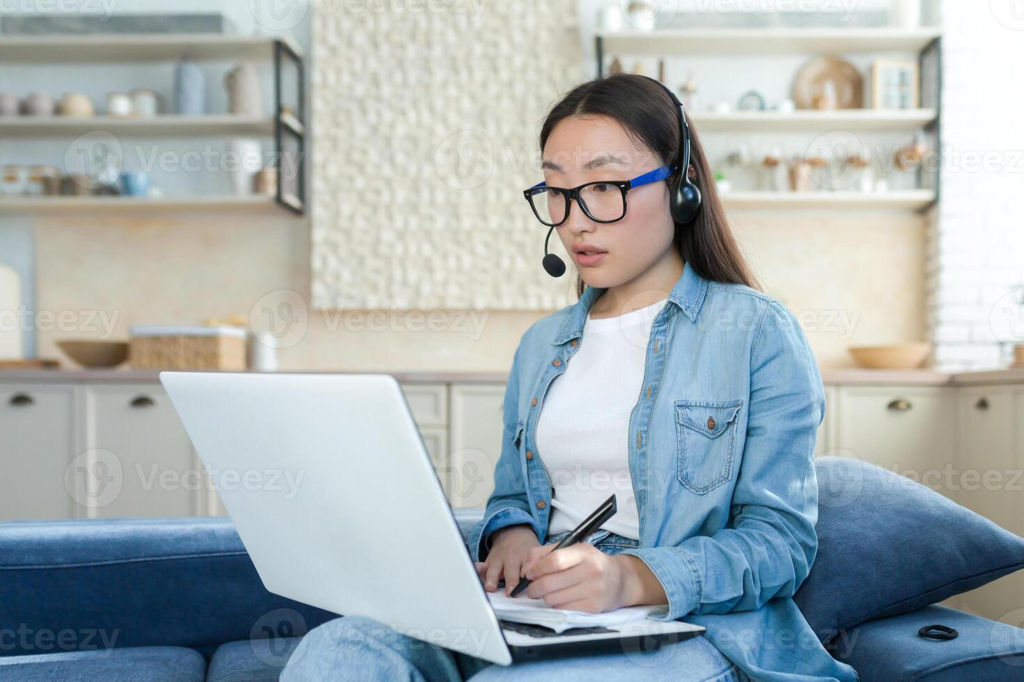 Work online. A young Asian woman is a call center operator. Sitting on the sofa at home wearing headphones with a microphone and a laptop, answering calls, consulting, writing down a request. photo