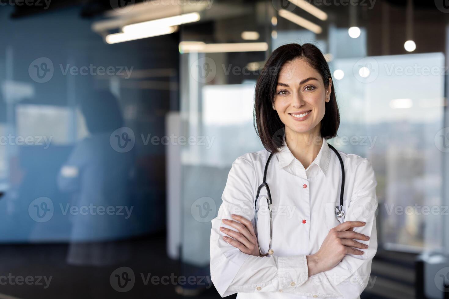 Professional female medical practitioner smiling confidently in a clinic setting, arms crossed, wearing a lab coat and stethoscope. photo