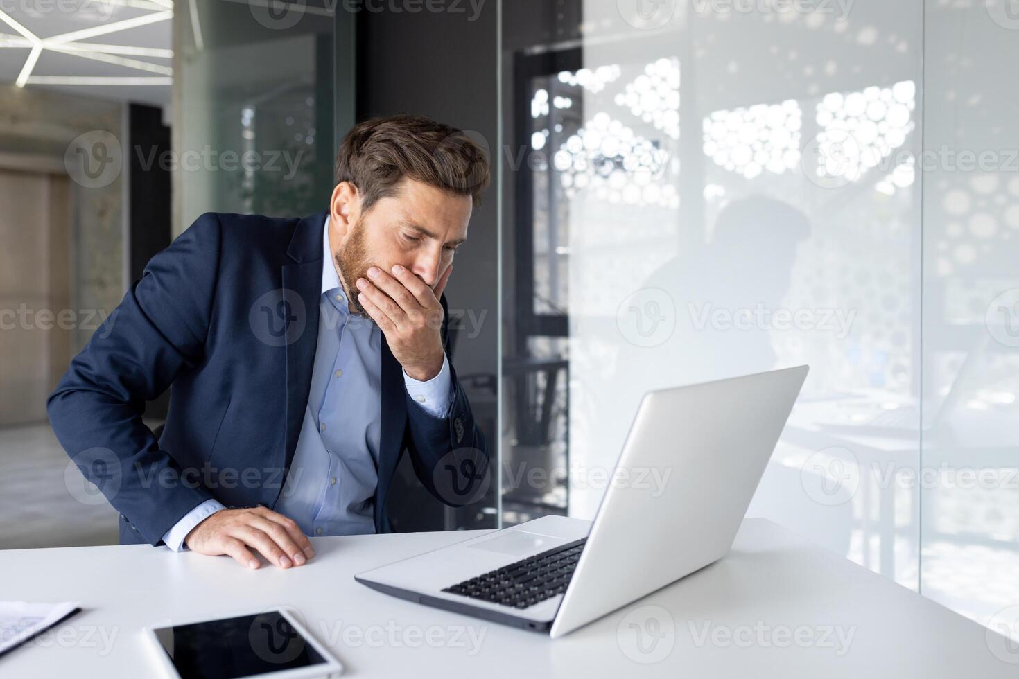 A young man businessman sits in the office at the work table and covers his mouth from nausea, he does not feel well. photo