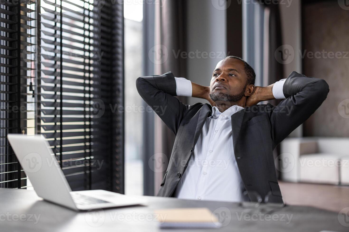 Overloaded black man in formal outfit leaning on back of chair while sitting with hands behind head by table with laptop. Tired lawyer stretching muscles and looking away during break from work. photo