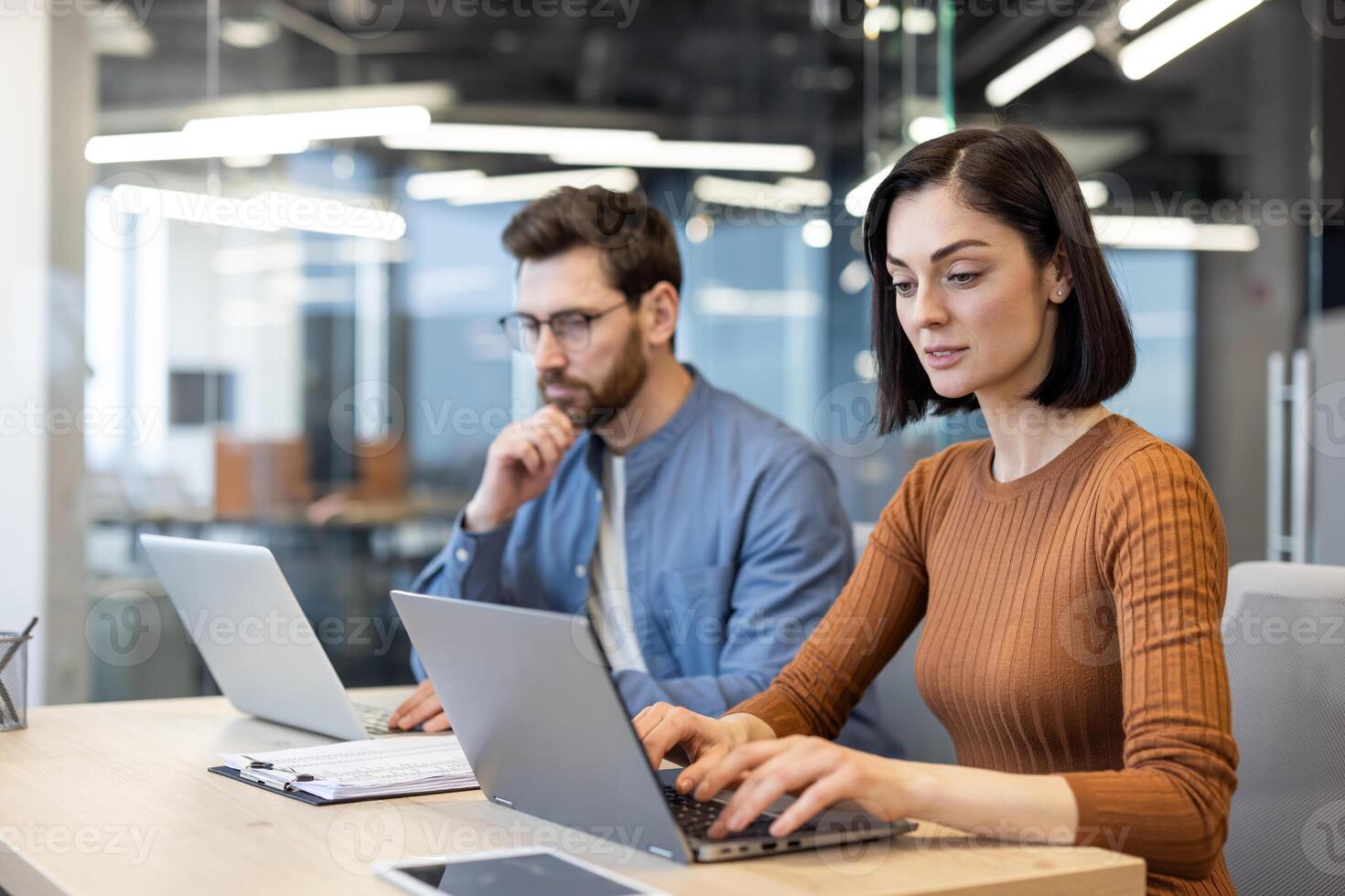 Caucasian woman sitting next to bearded man at shared workplace and typing on portable laptop. Ambitious colleagues working on personal results and solving list of tasks written on clipboard. photo