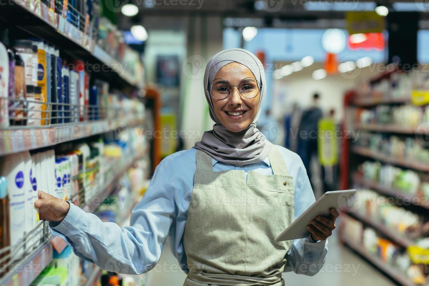 Portrait of a female saleswoman in a hijab, a salesperson in a household chemicals department is smiling and looking at the camera, holding a laptop tablet computer in her hands photo