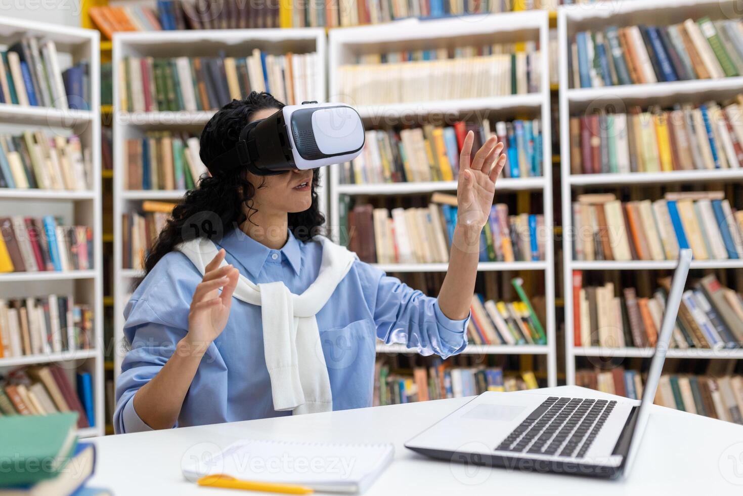 Young woman wears virtual reality headset in library, experiencing immersive technology with books and laptop around. photo