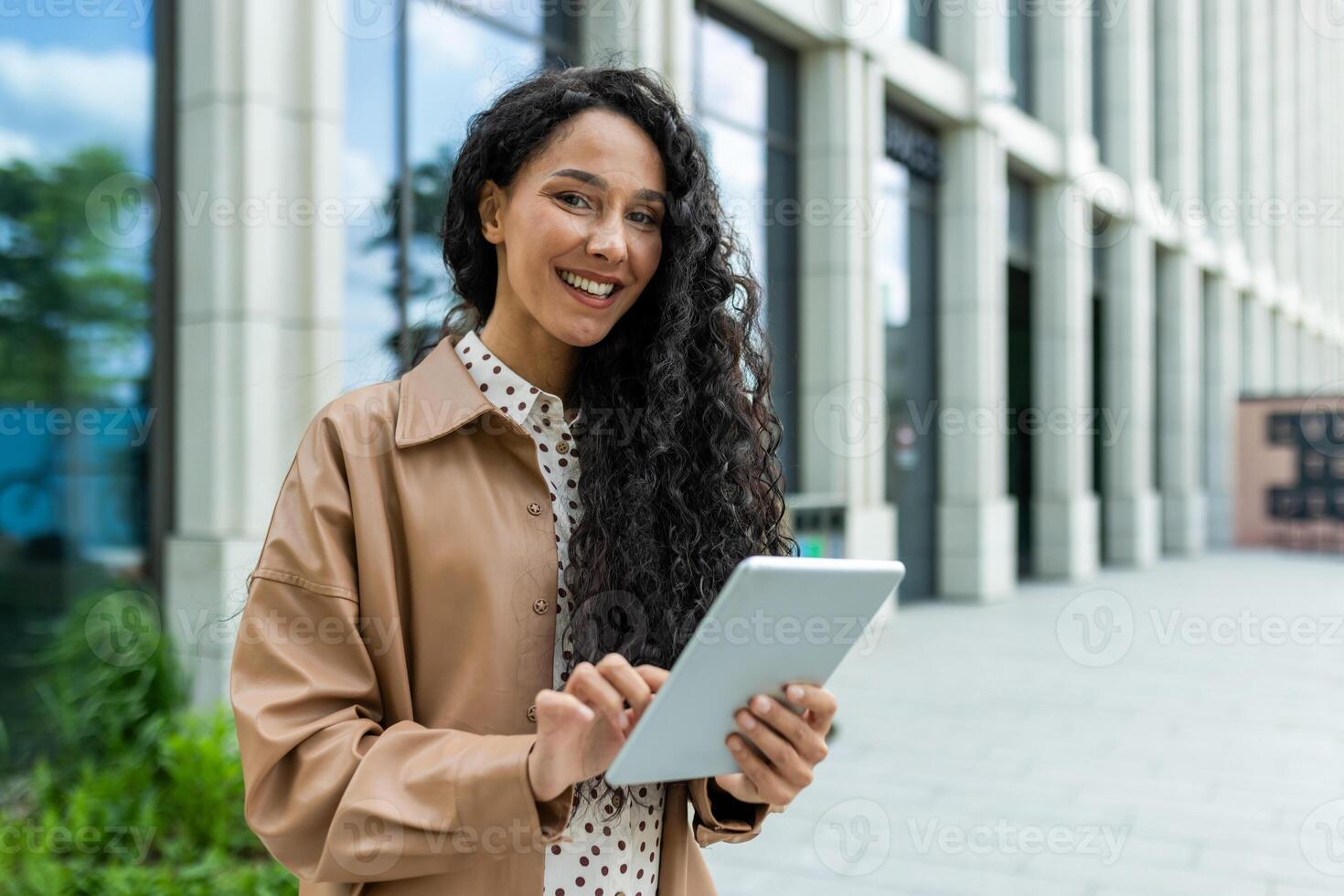 A professional woman stands outside an office building, engaged with a tablet. The scene suggests business, technology, and modern urban lifestyle. photo