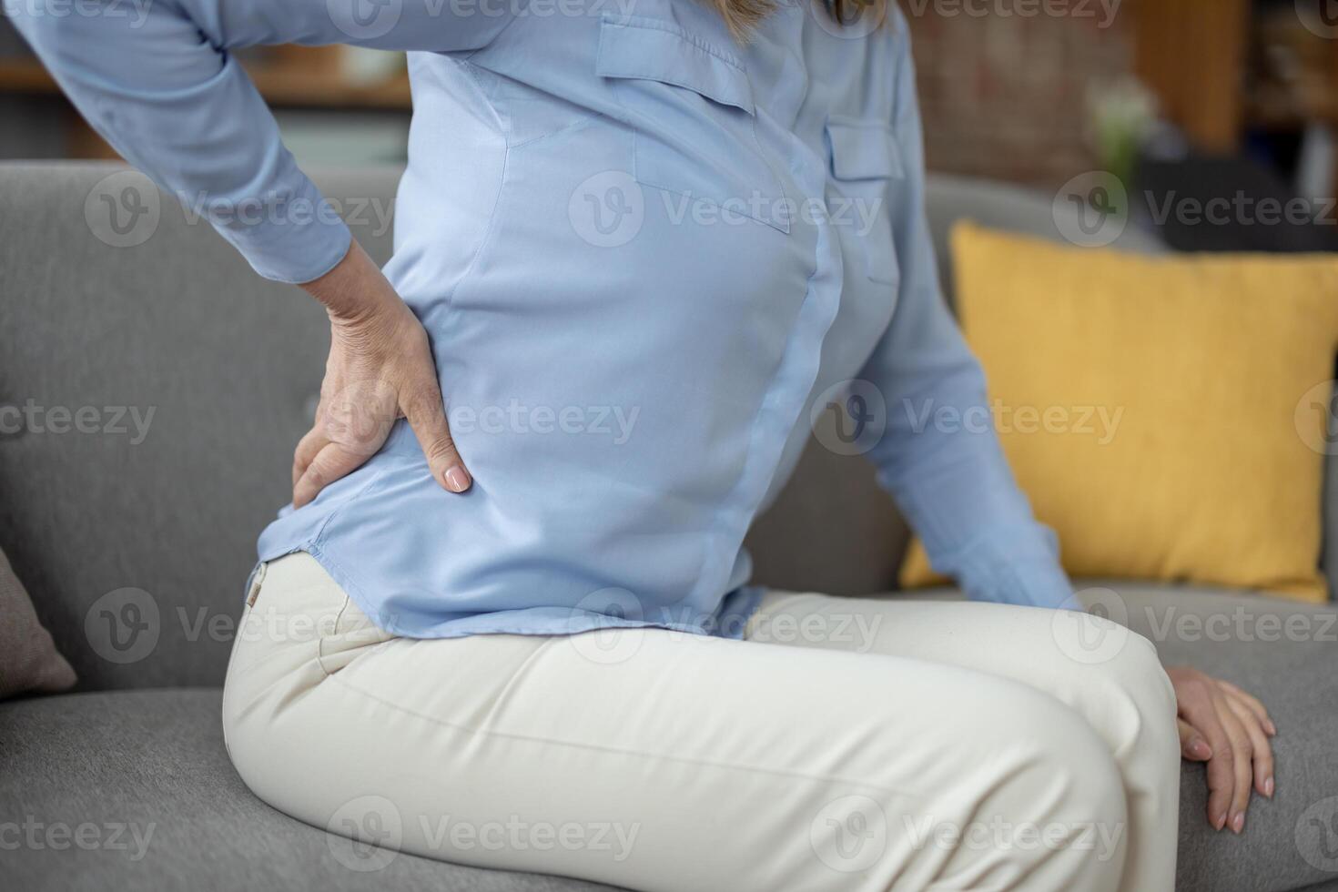 A senior female sitting on a sofa clutching her lower back in pain, highlighting the discomfort many elderly experience in daily life. photo