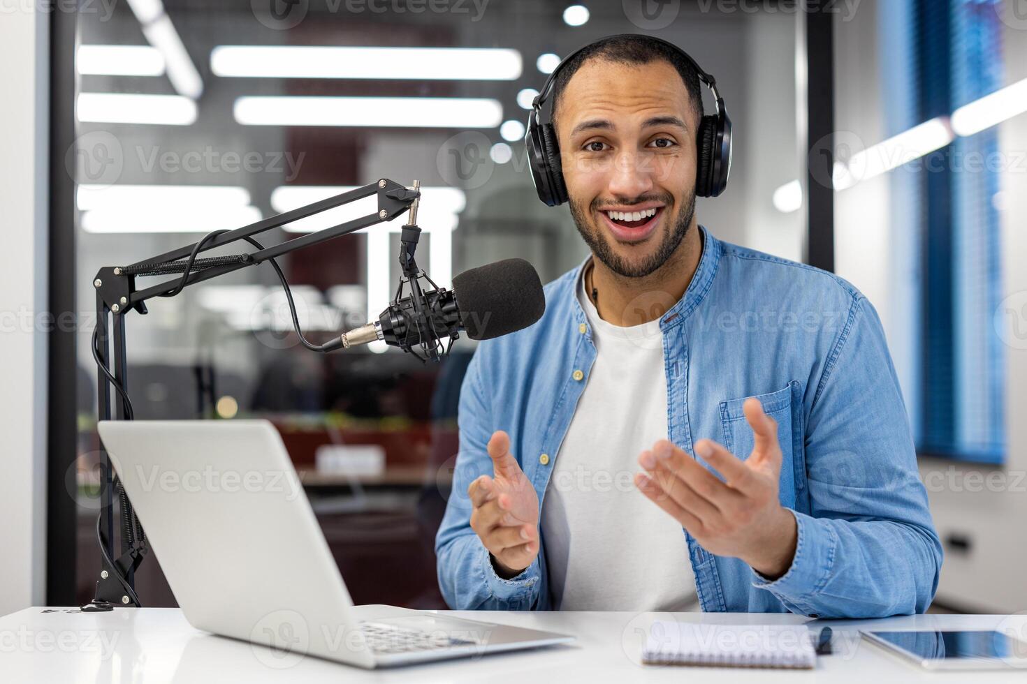 un indio hombre en casual atuendo es podcasting en un moderno vivo habitación, creando contenido con un profesional micrófono y ordenador portátil. foto