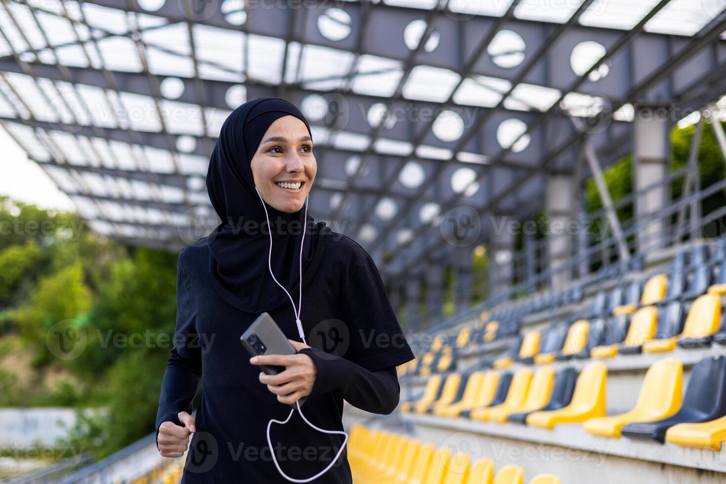 Young beautiful Muslim woman running in hijab in stadium, sportswoman smiling in headphones holding phone, listening to audio book. photo