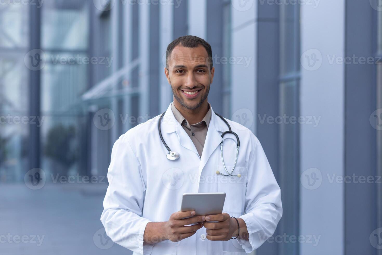 Portrait of happy and smiling hispanic doctor, man smiling and looking at camera, doctor using tablet computer for online consultation, outside of modern clinic photo