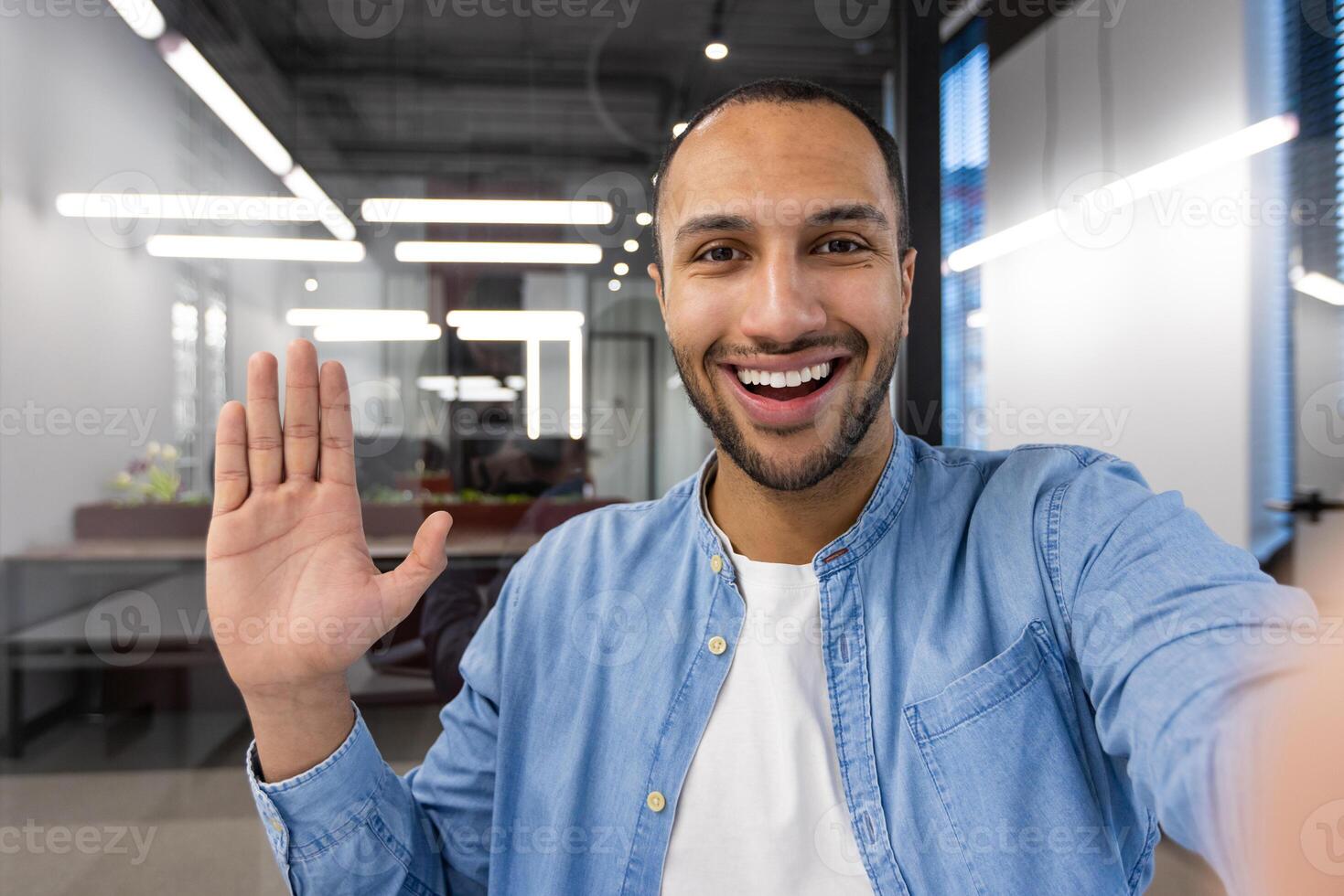 A man with a big smile on his face is waving at the camera. He is wearing a blue shirt and white shirt photo