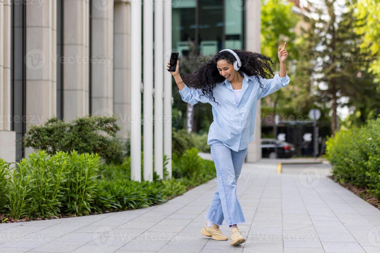 Young joyful beautiful woman dancing and singing in headphones while walking in the city, Hispanic woman with curly hair uses an application on the phone, to listen to music online. photo