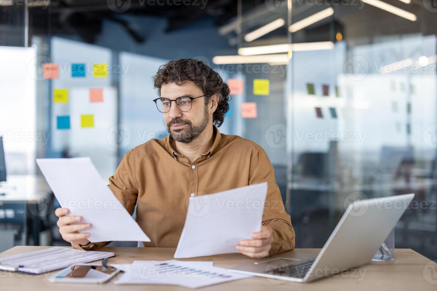 Thoughtful financial advisor in eyeglasses checking and analyzing printed graphs on paper sheets in personal workplace. Attentive man reading additional data for making deep research of investments. photo