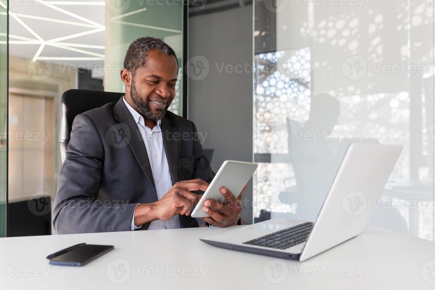 Mature adult african american boss investor at workplace using tablet computer, man happy with financial results and achievement smiling, using app inside office at table. photo
