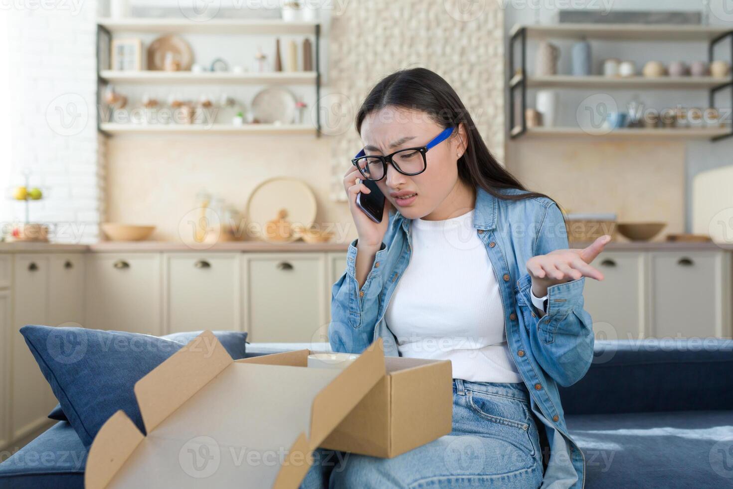 insatisfecho mujer recibido un roto paquete o empaquetar entregado desde un en línea en línea almacenar, asiático peleas con el cliente apoyo servicio, negociaciones a el operador en el teléfono, sentado hogar en el cocina foto