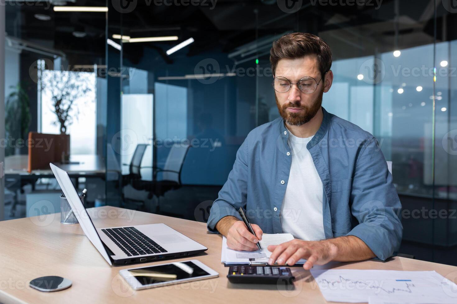 Serious and focused financier accountant on paper work inside office, mature man using calculator and laptop for calculating reports and summarizing accounts, businessman at work in casual clothes. photo
