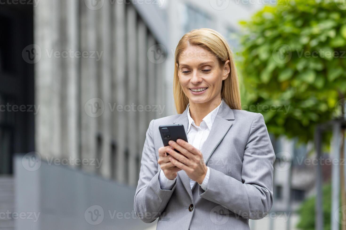 exitoso mujer jefe camina fuera de oficina edificio al aire libre, en negocio traje, mujer de negocios usos aplicación en teléfono, sonrisas contentamente lee Internet páginas, navega noticias, escribe texto mensaje, llamar. foto