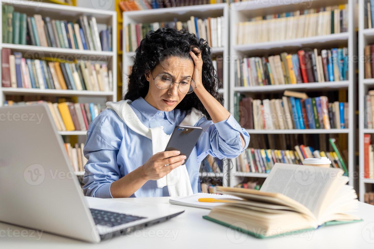 Anxious Hispanic student looking at phone with laptop and books at library desk. Concept of stress in education and learning difficulties. photo