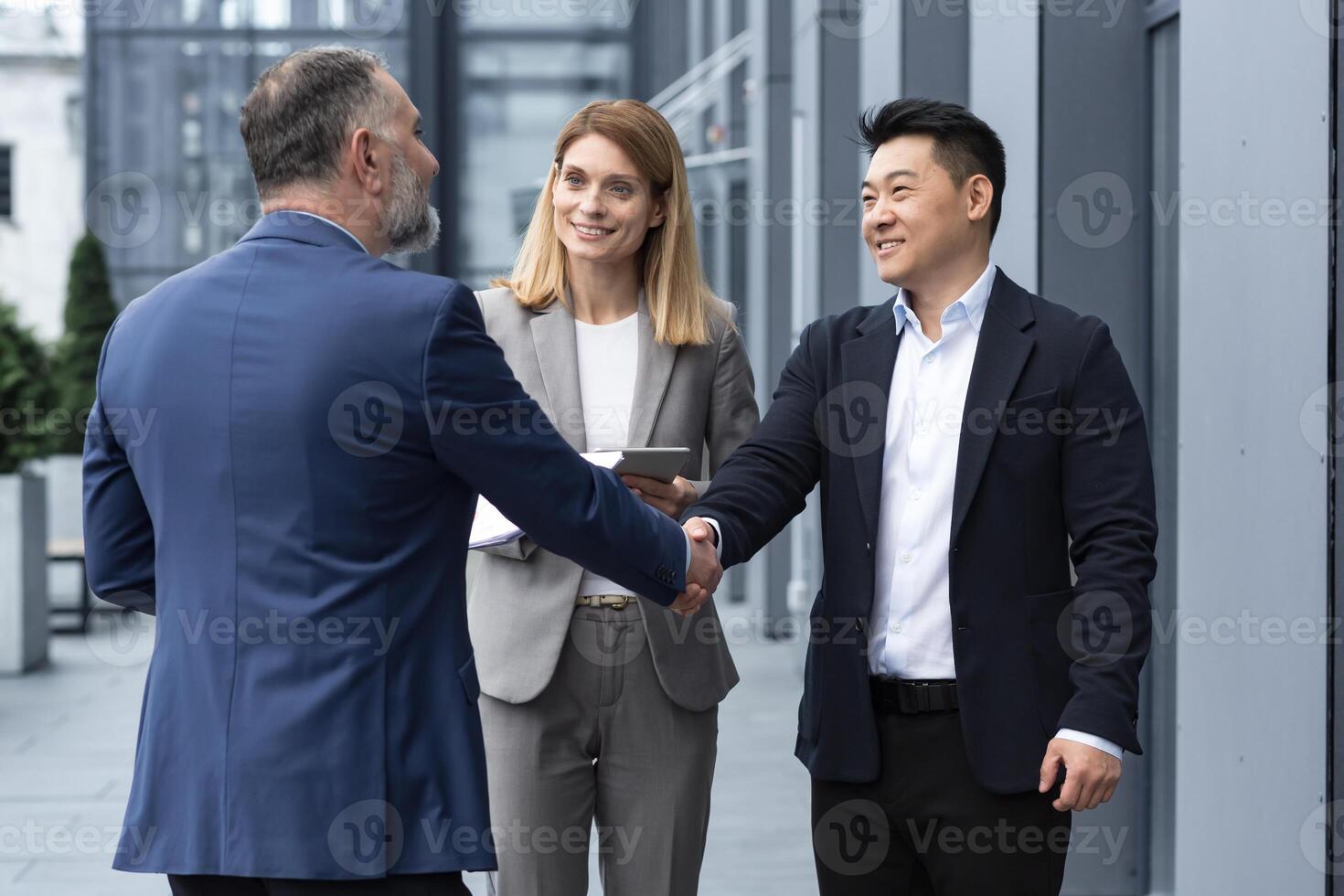 Meeting of three successful business people, diverse dream team man and woman outside office building, greeting and shaking hands, experienced professionals specialists in business suits talking photo