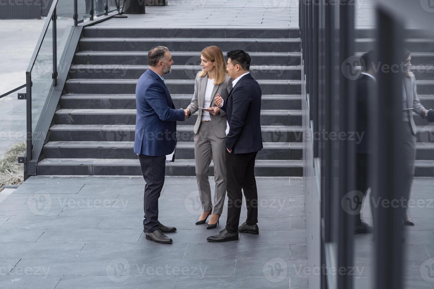 Meeting of three successful business people, diverse dream team man and woman outside office building, greeting and shaking hands, experienced professionals specialists in business suits talking photo