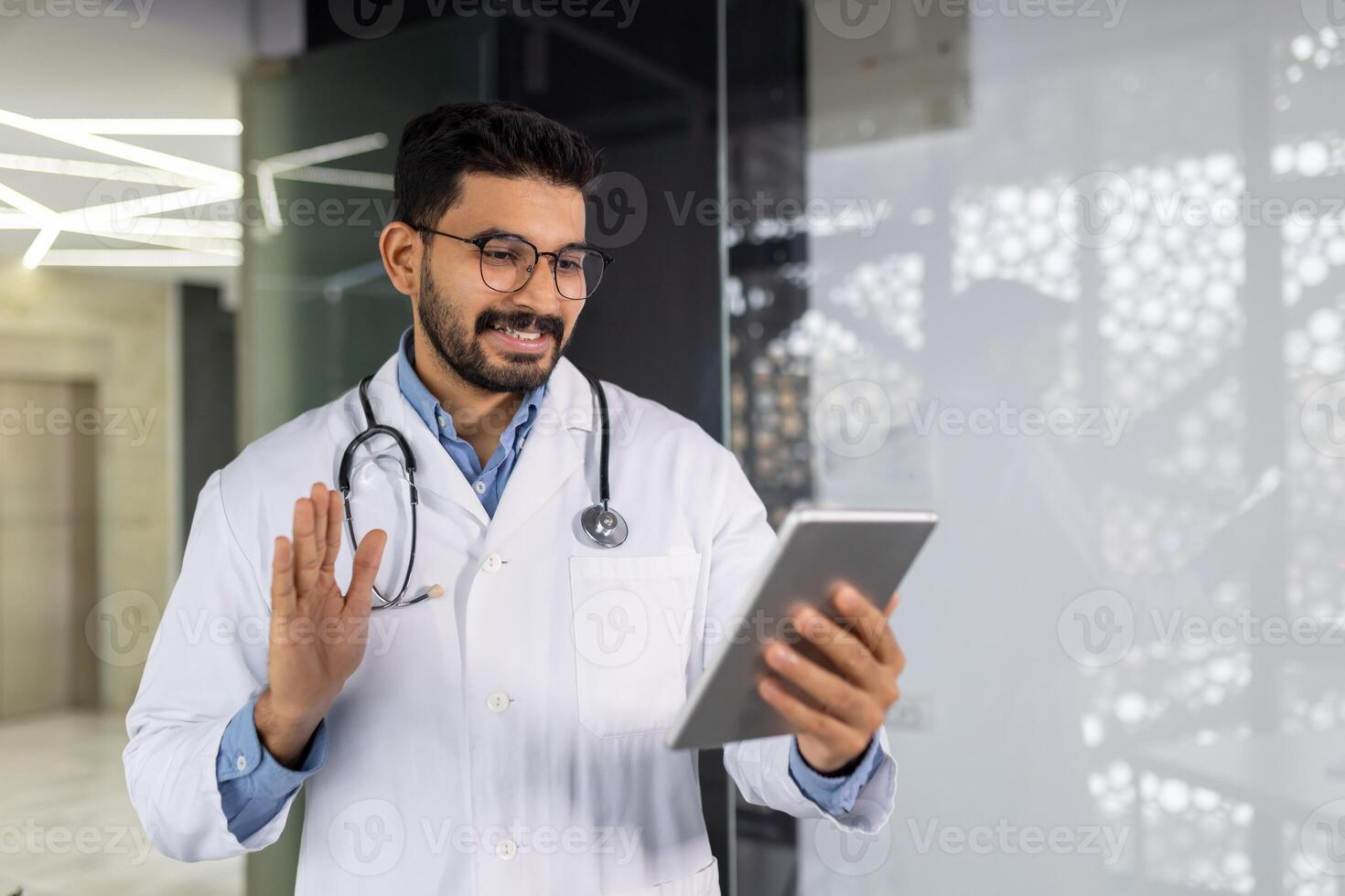 A young doctor smiles with a tablet computer in his hands, talks remotely with a patient, consults using a game, waves and greets, stands inside the clinic by the window. photo