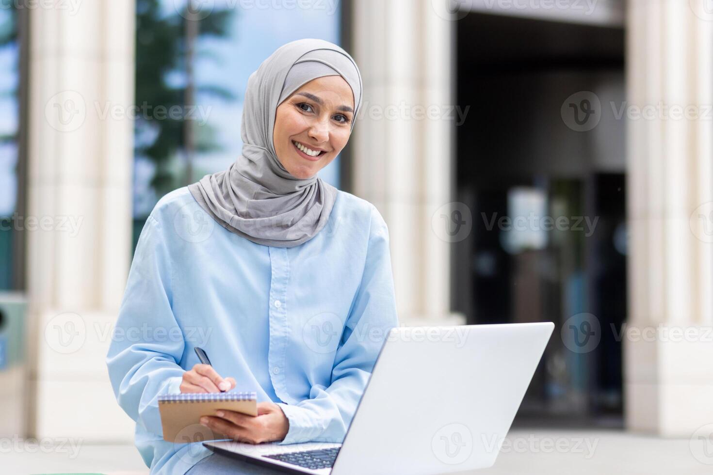 A young Muslim woman focused on studying outside, taking notes beside her laptop at a university campus. photo