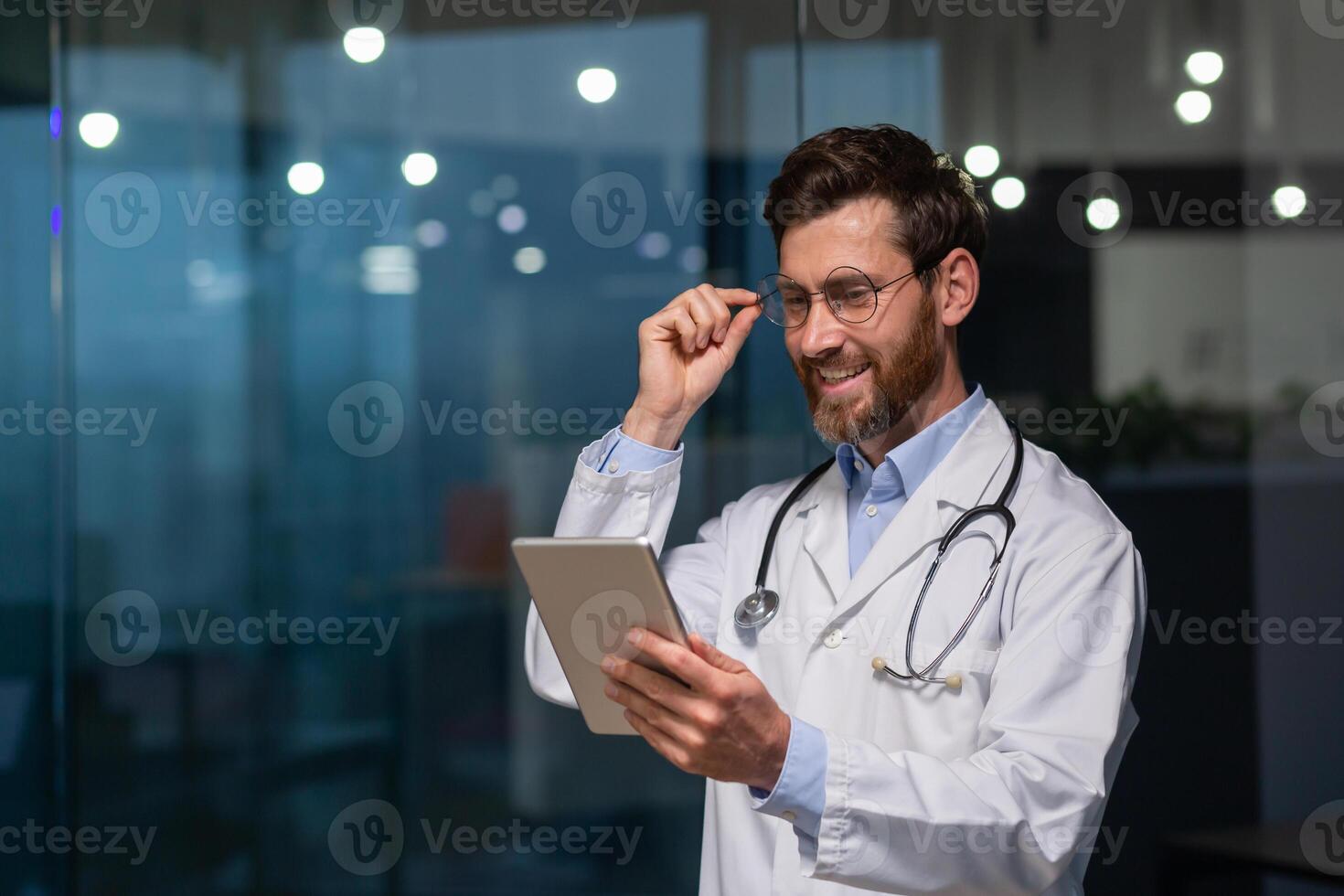 A young male doctor, intern, intern is standing in an office in a hospital in a white coat and with a stethoscope. He works on a tablet, holds his glasses in his hand, and smiles. photo