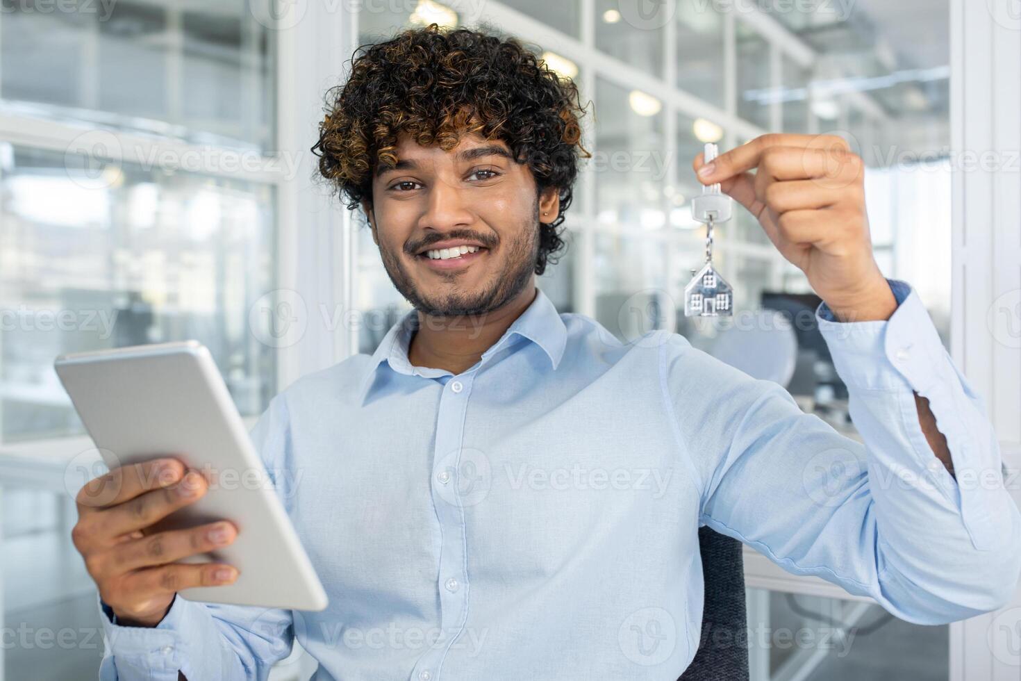 A cheerful young man showcases house keys while using a tablet in a light-filled office. Perfect image conveying success in real estate and technology. photo