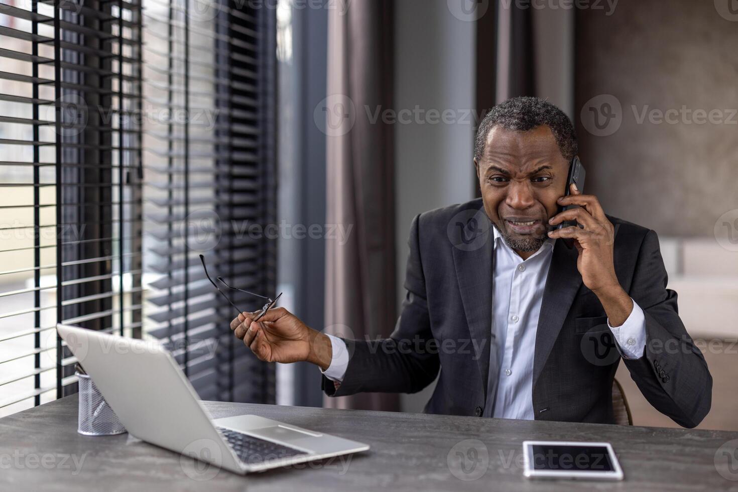 Furious diverse male in formalwear holding eyeglasses while having unpleasant phone conversation. Angry distant worker hearing bad breaking news while sitting by desktop with portable computer. photo