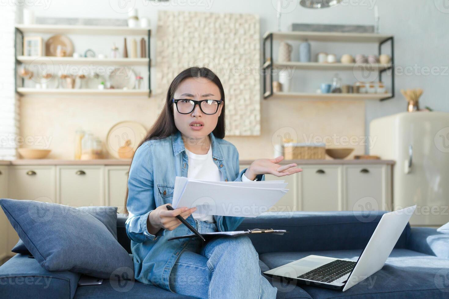Confused and nervous young Asian woman sitting on sofa at home and working with documents and laptop. Checks the accounts and finances of the family budget. He looks at the camera, spreads his hands. photo