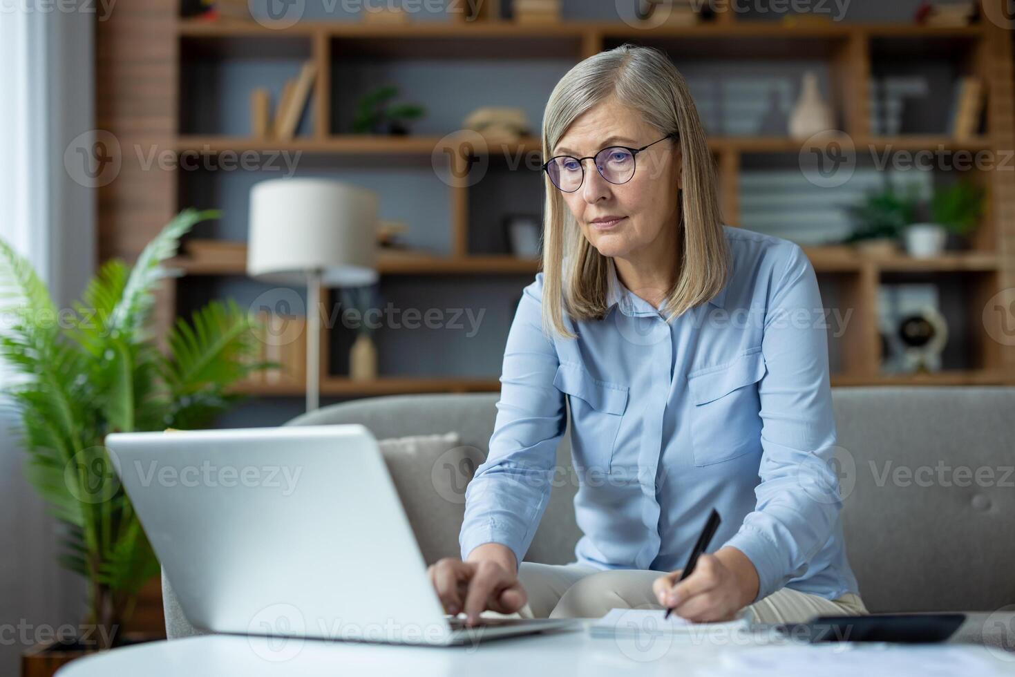 Stylish mature woman sitting on couch and using portable computer while writing in notebook at home. Attentive female joining remote classes for advanced training and making remarks from lecturer. photo