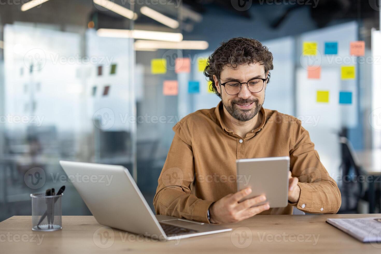 Casually dressed businessman working with a digital tablet and laptop at a wooden desk in a contemporary office setting. Productive and relaxed atmosphere. photo
