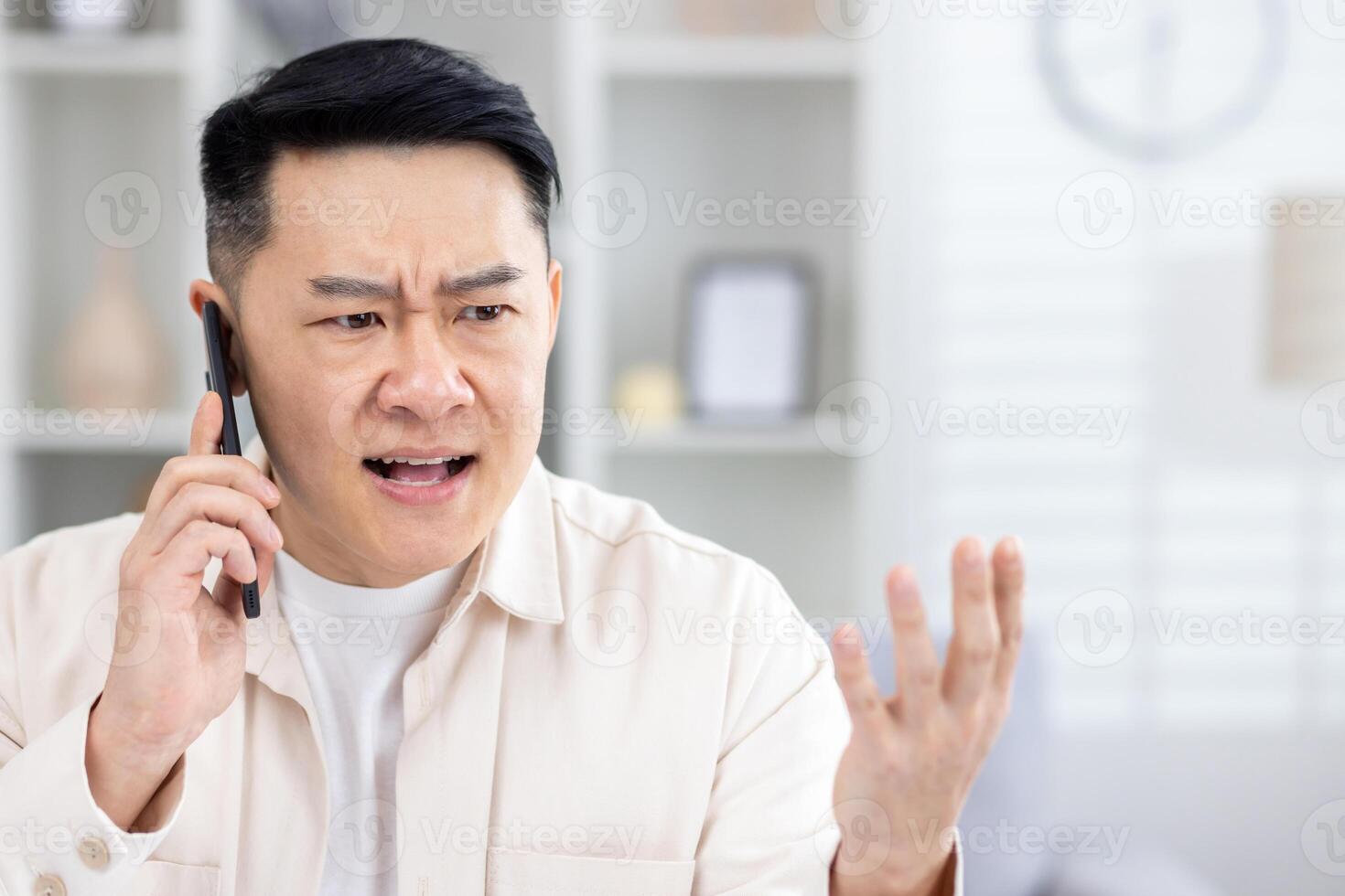 Angry angry and upset man talking on phone at home, asian working remotely from home office sitting at table in living room, freelance businessman at work. photo