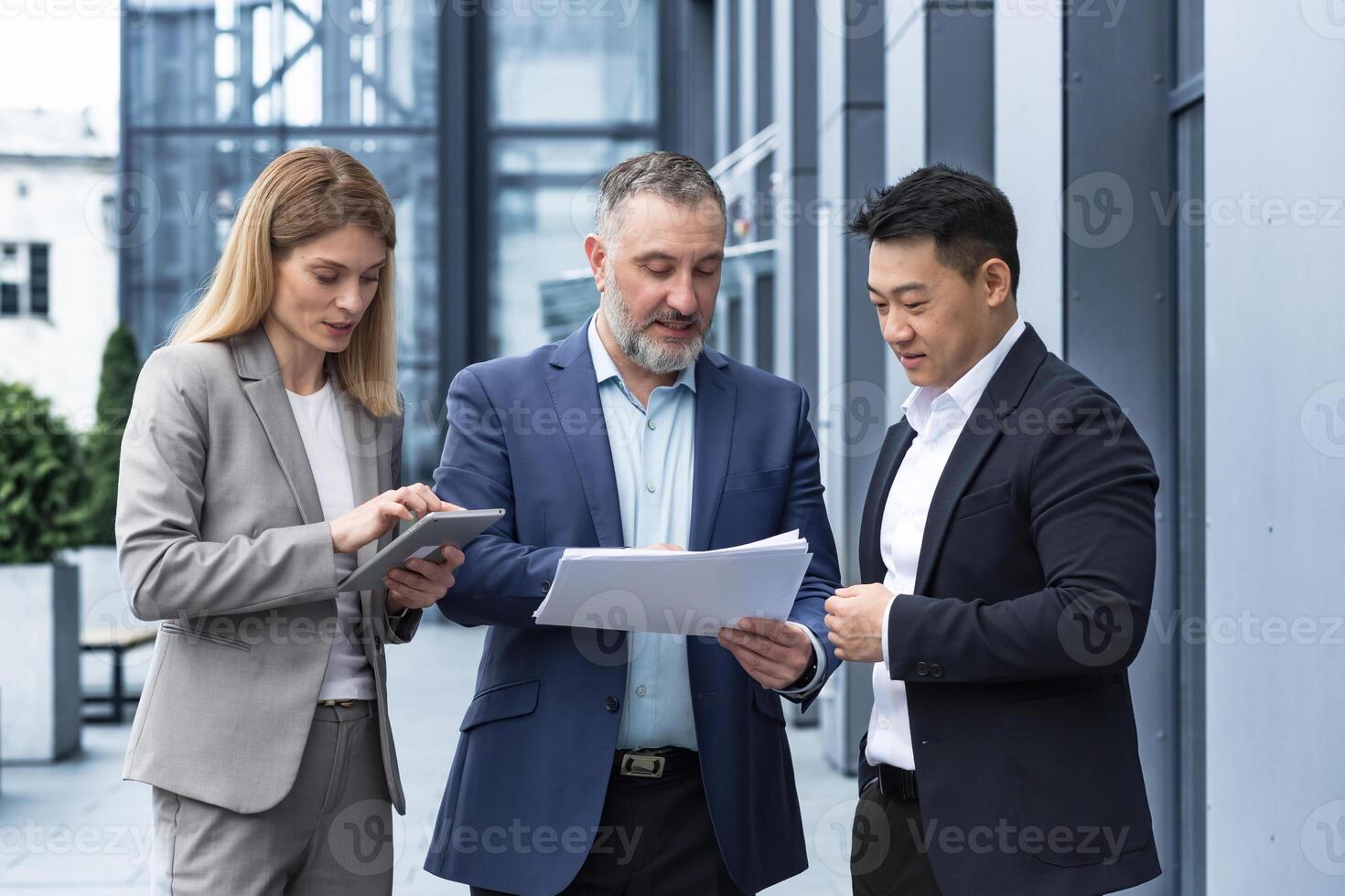 International business team meeting mature man, Caucasian woman, Asian employee communicate . group of entrepreneurs in formal suits discuss a project document near office center on street photo